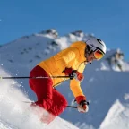 Skier in mid-descent on a snow-covered mountain slope with dramatic alpine peaks and pine forest below on a bright clear day