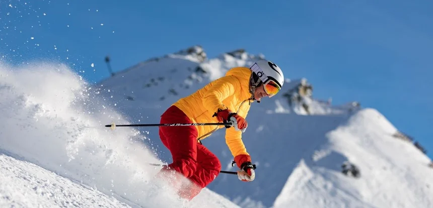 Skier in mid-descent on a snow-covered mountain slope with dramatic alpine peaks and pine forest below on a bright clear day