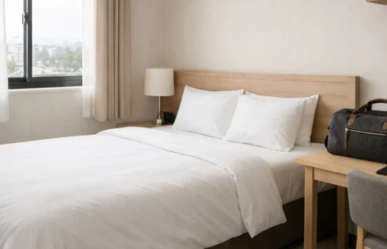 Clean minimalist budget hotel room with natural light through a large window and a travel bag on the desk