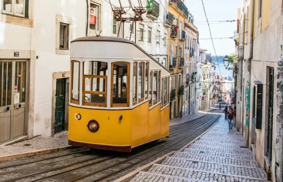 A yellow Lisbon tram climbing a steep street lined with colorful Portuguese tiled building facades and a traveler on the sidewalk in warm afternoon light