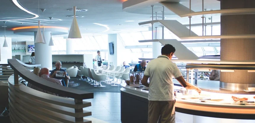 Traveler relaxing in a plush seat in a calm airport lounge with a buffet food spread and drinks bar visible