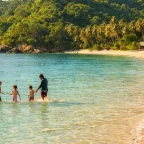 Family splashing at the shoreline of a calm turquoise tropical beach with lush green hills in the background